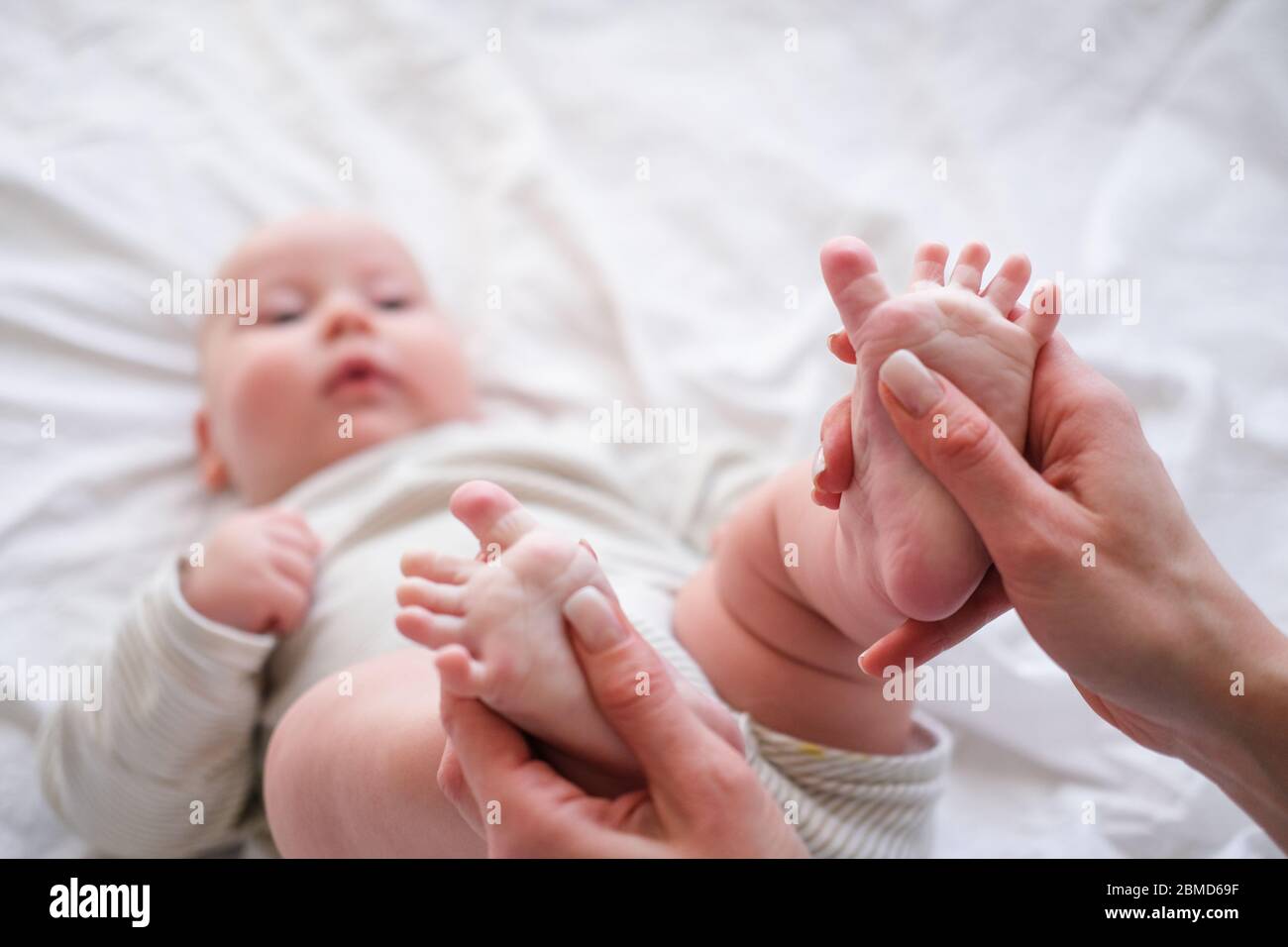 Babynahrung in den Händen der Mutter. Die junge kaukasische Frau massiert zu Hause für glückliches Säuglingsbaby auf weißem Bett. Babypflege, Sport und glückliche Mutterschaft. Stockfoto