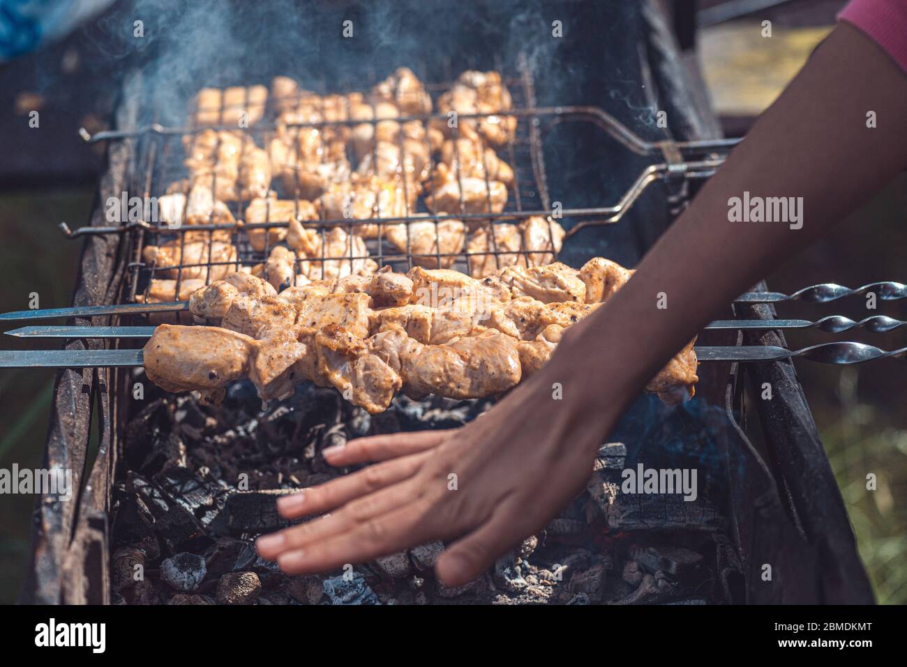 Die Hand einer Frau überprüft den Grad der Bereitschaft eines Grills, der auf Kohlen unter den Strahlen der Sommersonne gekocht wird Stockfoto