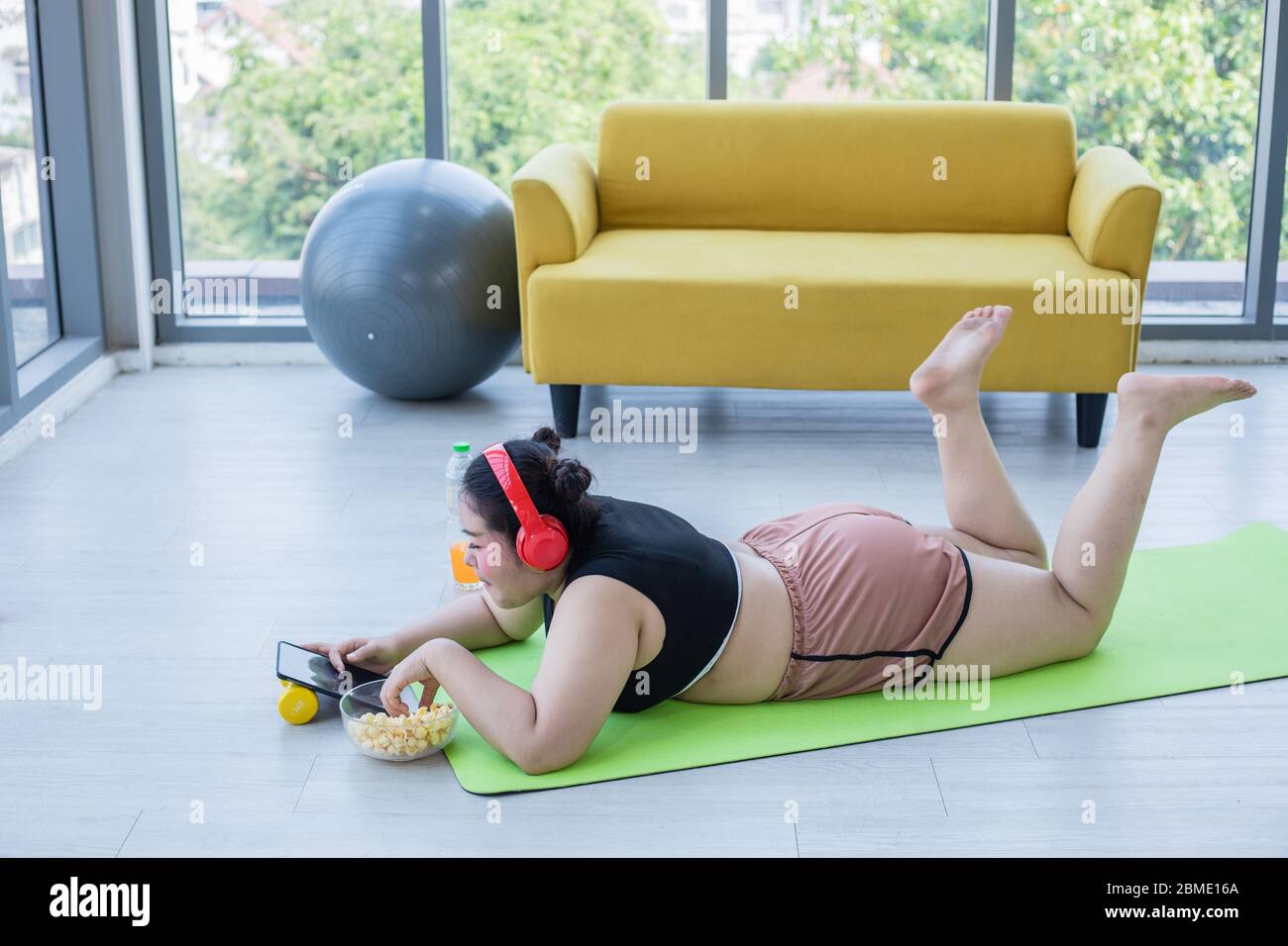 Asiatische übergewichtige Frau mit Tablette und. Tragen Kopfhörer für Musik hören und sie ist die Ausübung zu Hause, asiatische Mädchen genießen Essen und Popcorn Stockfoto