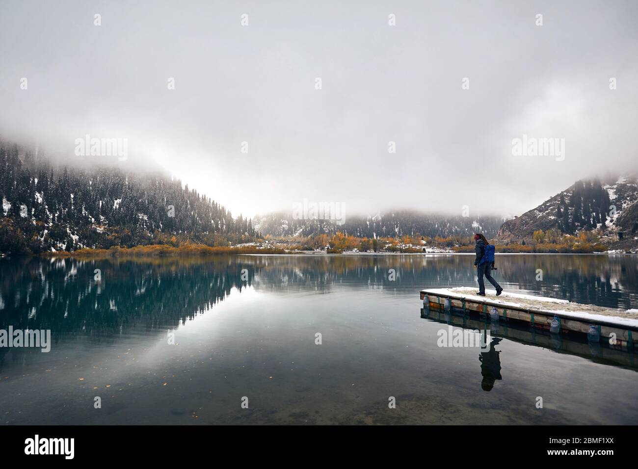 Einsamer Mann mit Rucksack und Kamera zu Fuß den Pier am nebligen See mit Reflexion an der Herbst in Kasachstan und Zentralasien Stockfoto