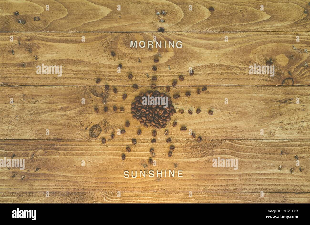 Kaffeebohnen, die wie die Sonne auf einem hölzernen Hintergrund geformt sind, mit den Wörtern „Morgensonne“, die mit hölzernen Buchstaben geschrieben werden Stockfoto