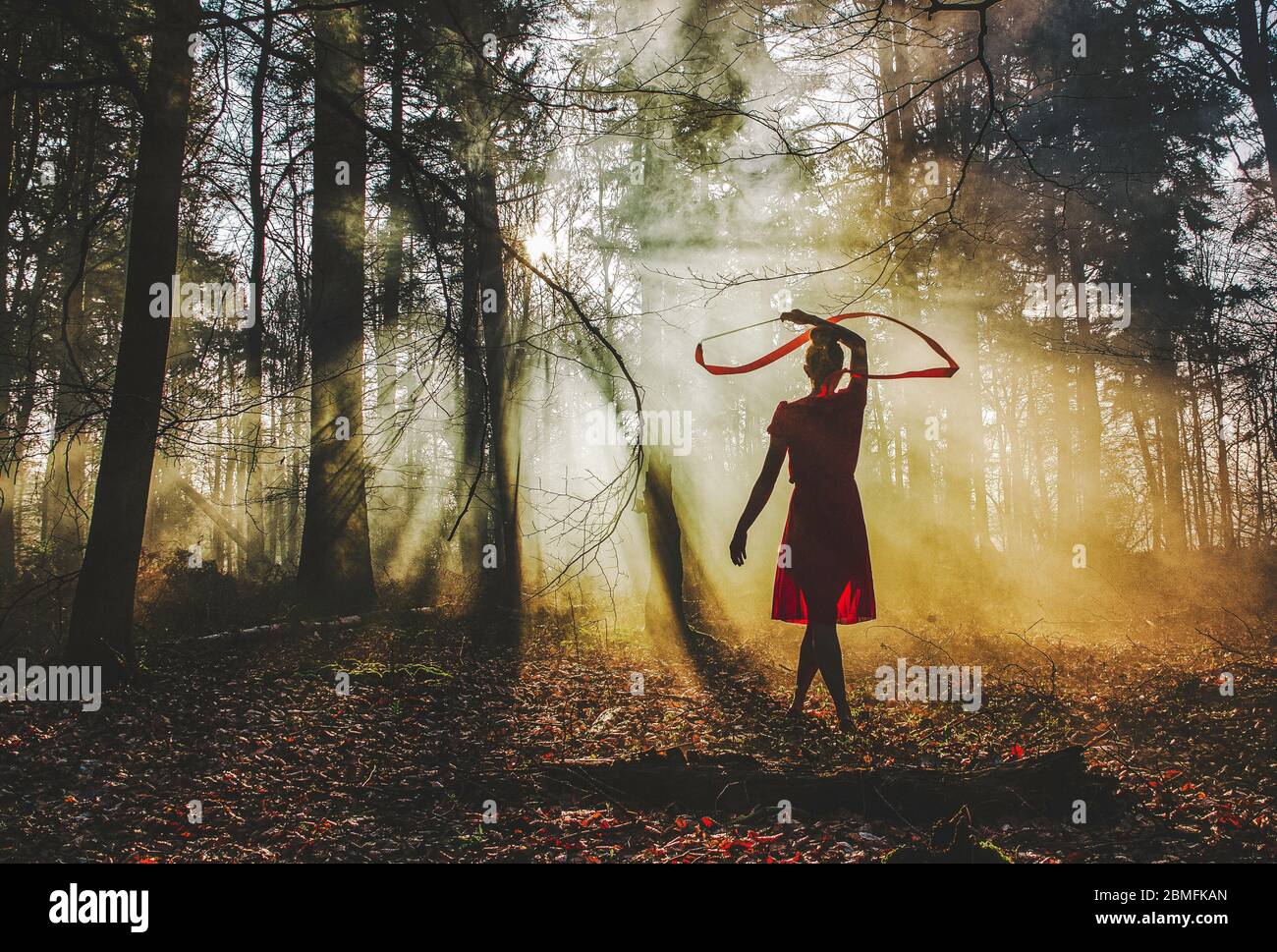 Eine Frau, die in einem Wald mit einem roten Band tanzt, während sie ein rotes Kleid trägt, während Sonnenstrahlen durch die Bäume scheinen Stockfoto