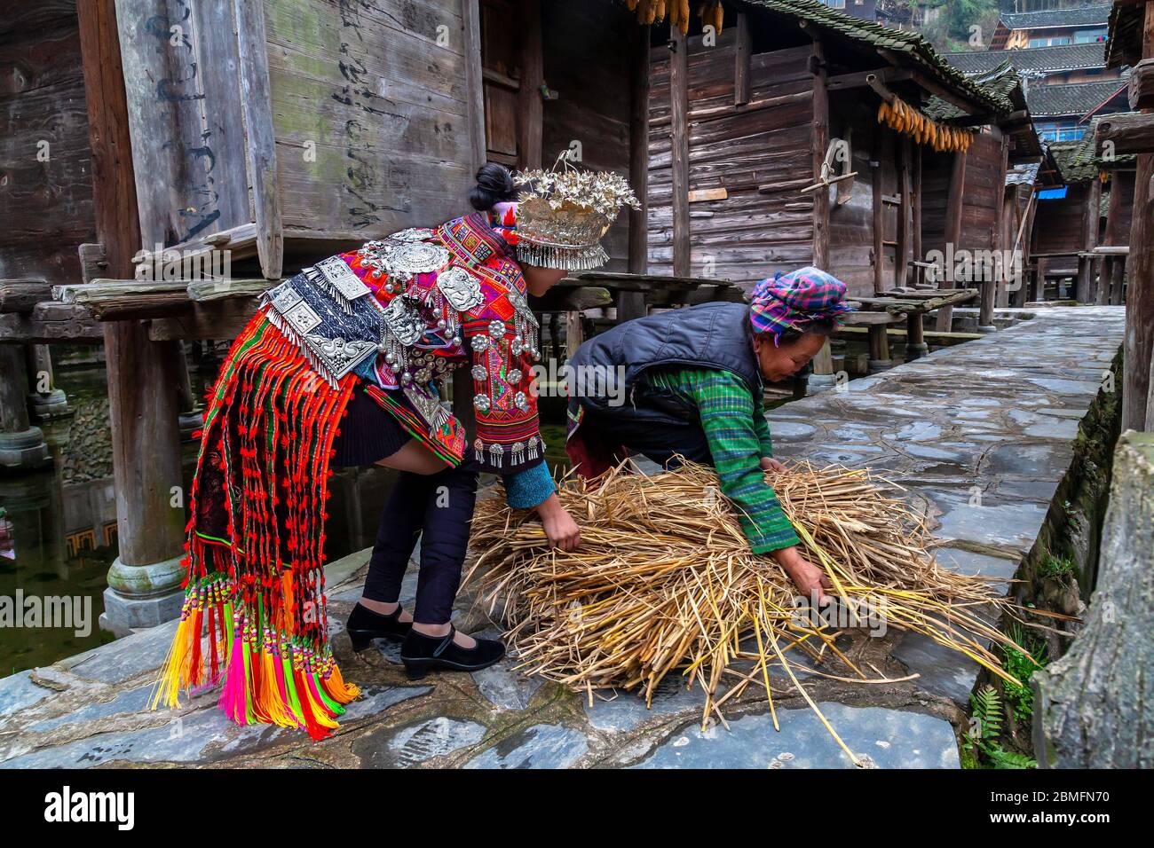Eine junge Frau in einem traditionellen Kleid, die einer älteren Frau mit ihrer Ladung Heu hilft.Sie gehören zu den Datang kurzen Rock Miao Minderheitsmenschen. Kail Stockfoto