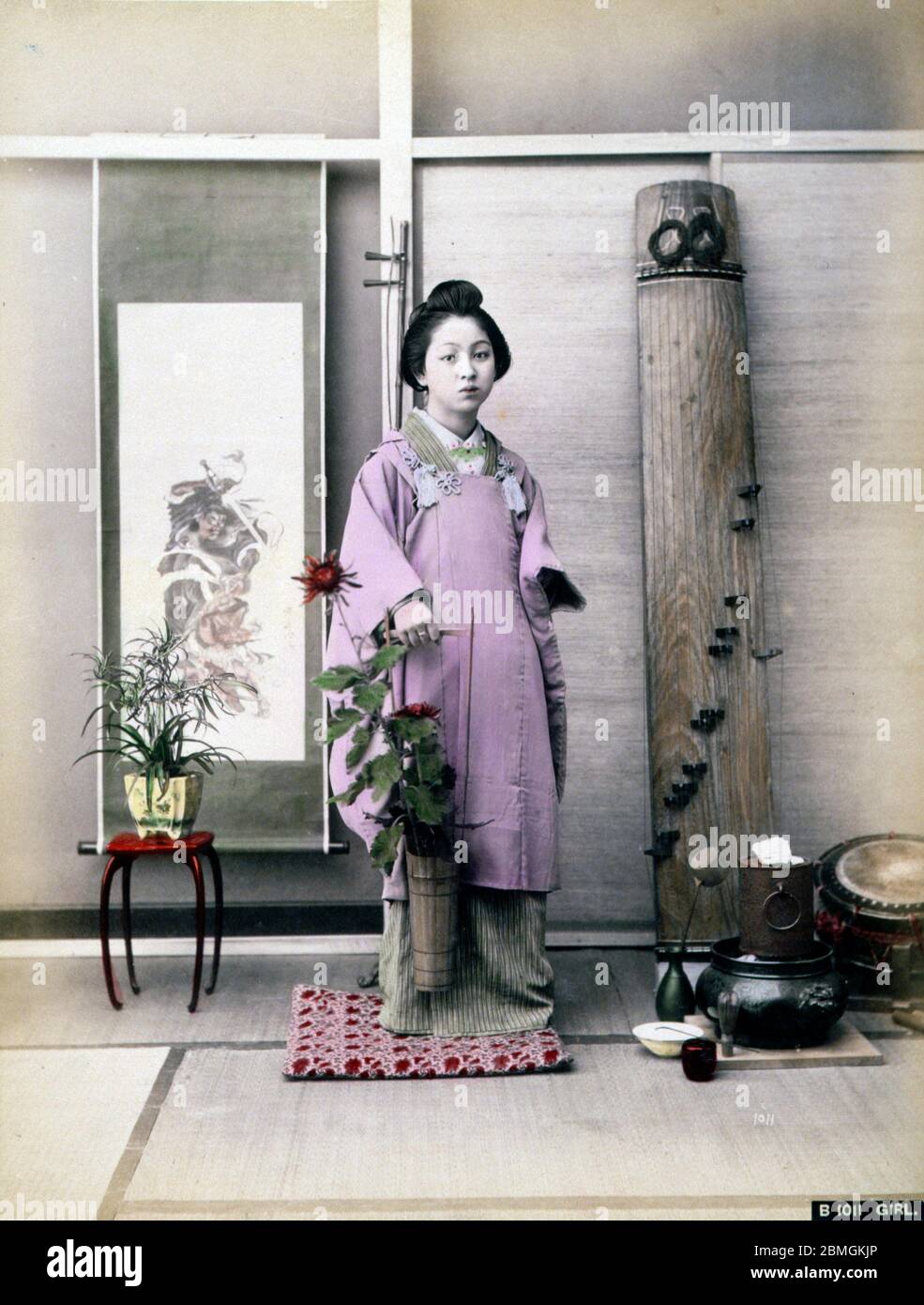 [ 1880er Japan - Japanische Frau mit Blumen ] - EINE Frau im Kimono, die eine Bambusvase mit Blumen hält. Ein Hibachi-Brazier, eine Taiko-Trommel, die als Ootsutsumi bekannt ist, und eine Koto, eine Art Harfe, sind auf der Rückseite sichtbar. Vintage Albumin-Fotografie aus dem 19. Jahrhundert. Stockfoto