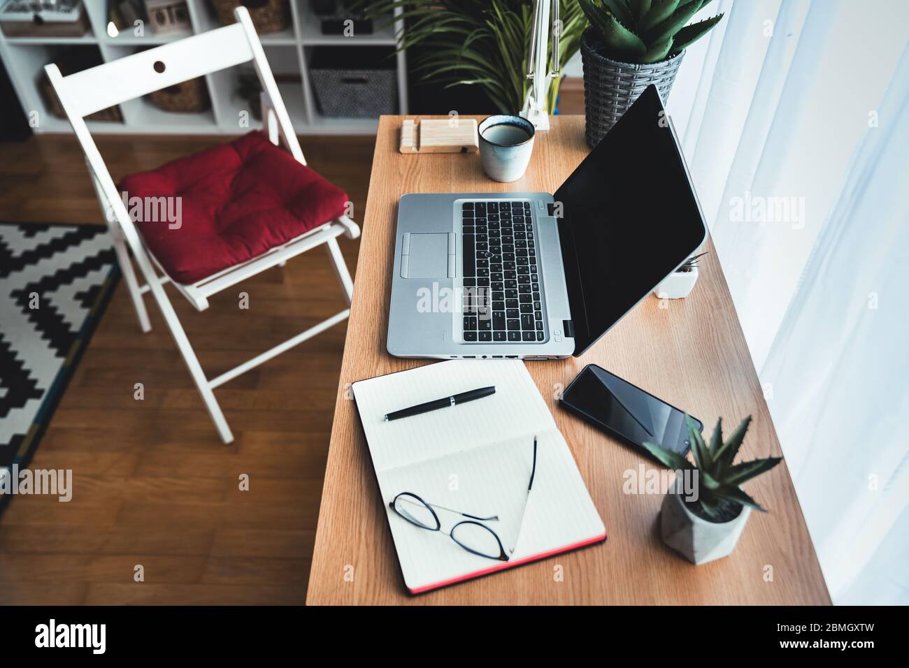 Draufsicht auf einen Schreibtisch mit Laptop in einem kreativen Home Office. Auf dem Tisch liegend: Smartphone, Notebook, Kaffeebecher, Stift, Augengläser, grüne Pflanzen. Stockfoto