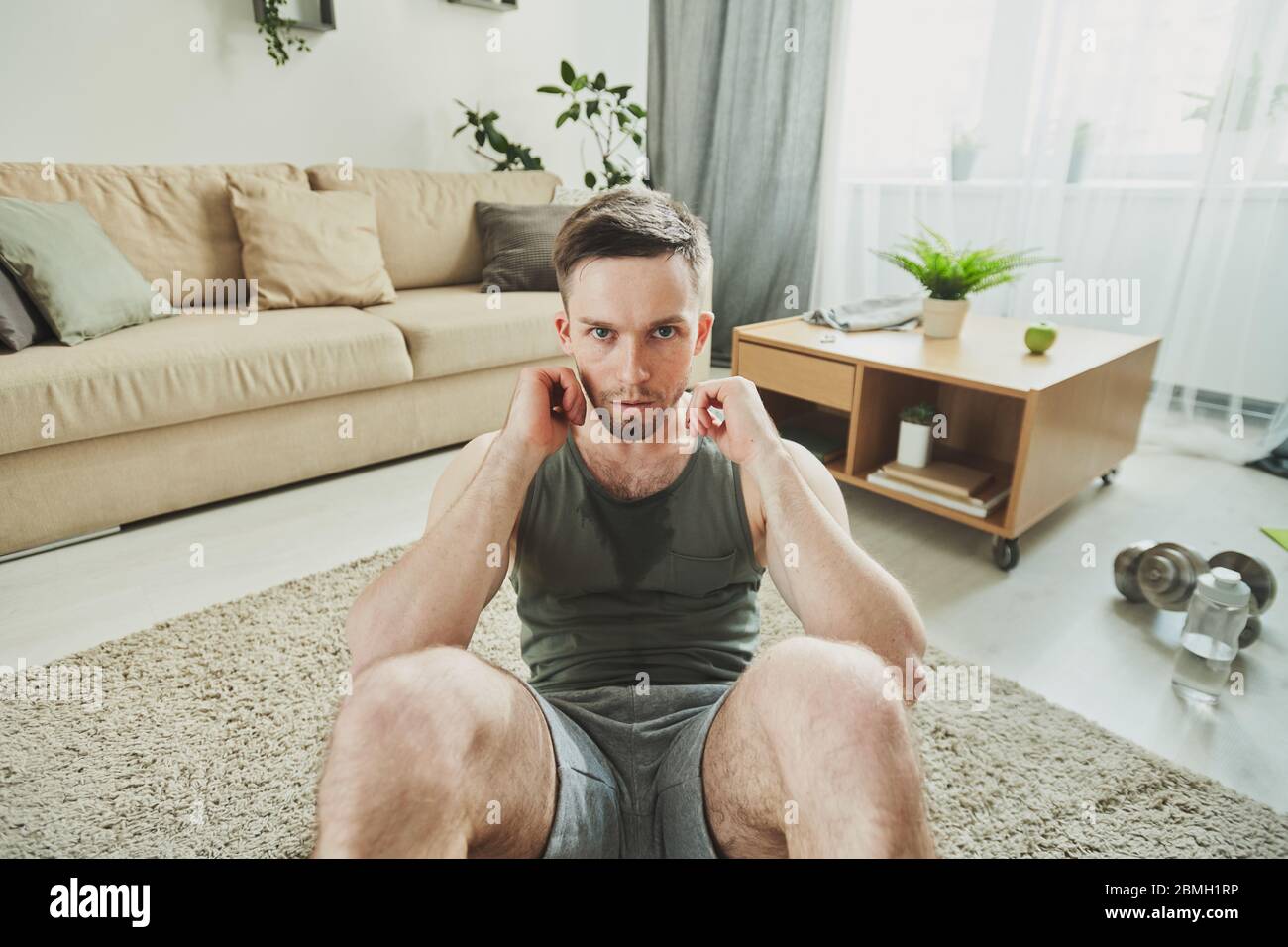 Junge ernsthafte schweißtreibende Athlet macht Sit-ups vor der Kamera und schaut Sie an, während Sie auf dem Teppich in der häuslichen Umgebung sitzen Stockfoto