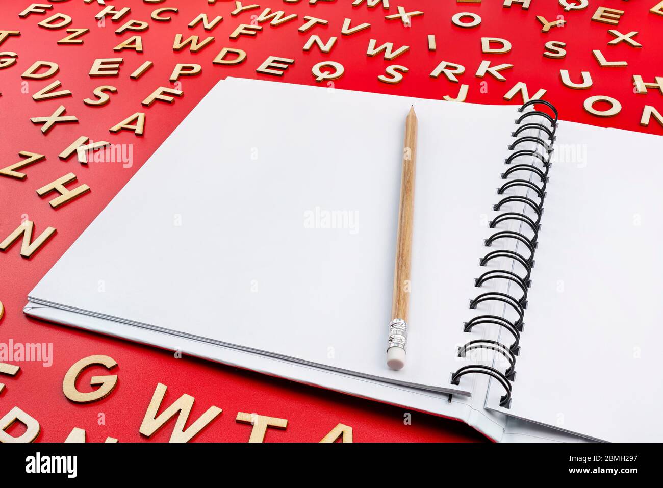 Ein offenes Notizbuch mit weißen Seiten und einem Holzstift auf rotem Grund mit verstreuten Buchstaben aus Holz. Stockfoto
