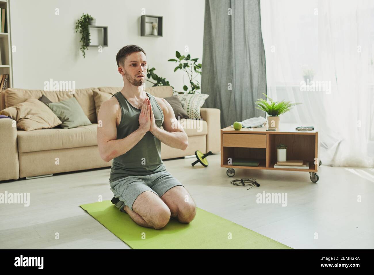 Ruhiger junger Mann in der aktiven Haltung auf den Knien mit Händen zusammen nahe an der Brust beim Üben Yoga oder Entspannungsübungen Stockfoto
