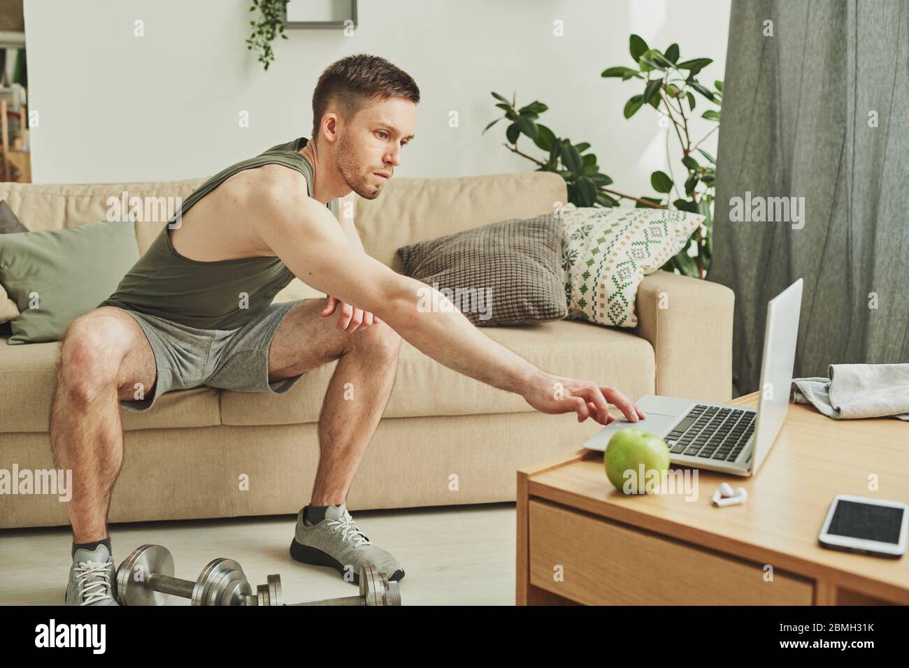 Junger Sportler in activewear sitzt auf der Couch, während Stretching Arm zu Laptop, um die Start-Taste drücken und sehen Sie Online-Video des Trainings Stockfoto