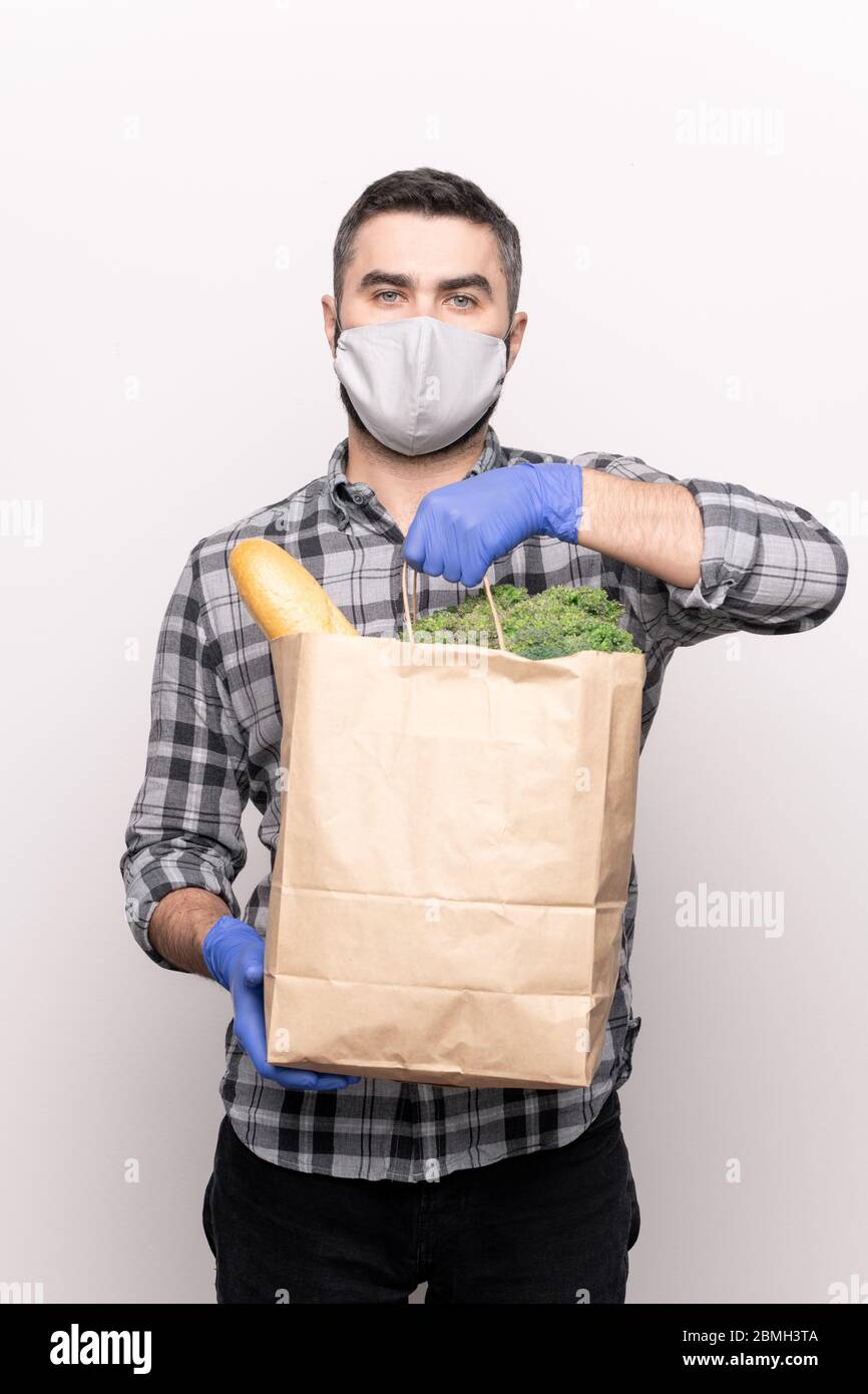 Seriöse Kurier in Schutzmaske und Handschuhe liefern frische Lebensmittel zu Ihnen nach Hause und halten Papierpaket mit Brot und Gemüse Stockfoto