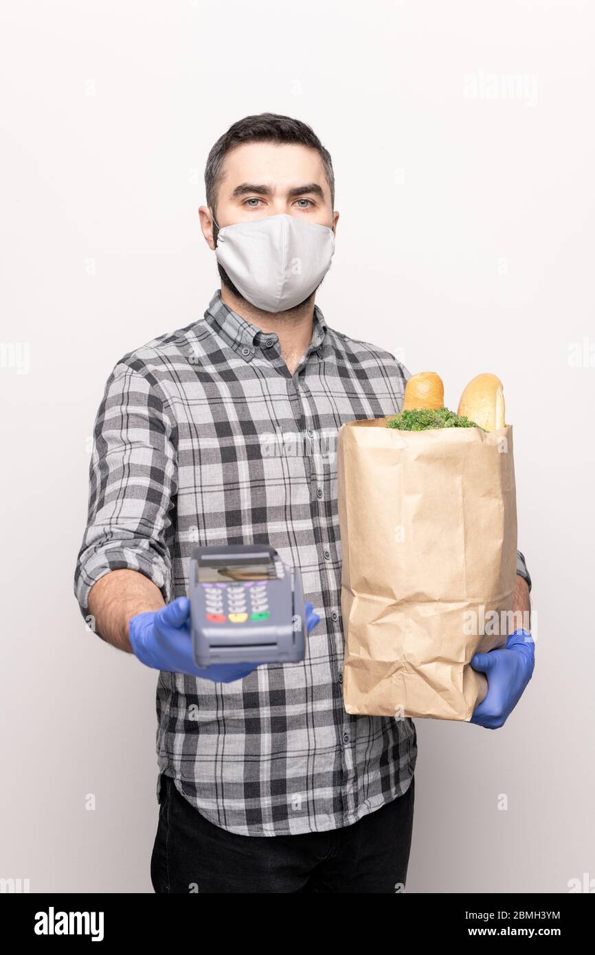 Junge Kurier in Hemd und Schutzhandschuhe und Maske hält Zahlungsmaschine und Papiertüte mit bestellten frischen Lebensmitteln Stockfoto