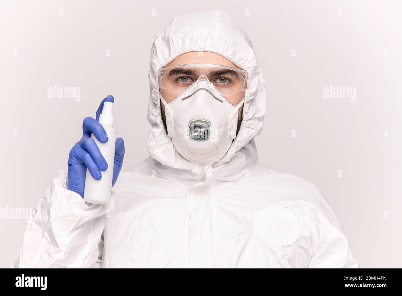 Schöner junger Mann in Schutzhüllen, Atemschutzmaske, Handschuhen und Brillen, die Desinfektionsmittel isoliert um sich herum sprühen Stockfoto