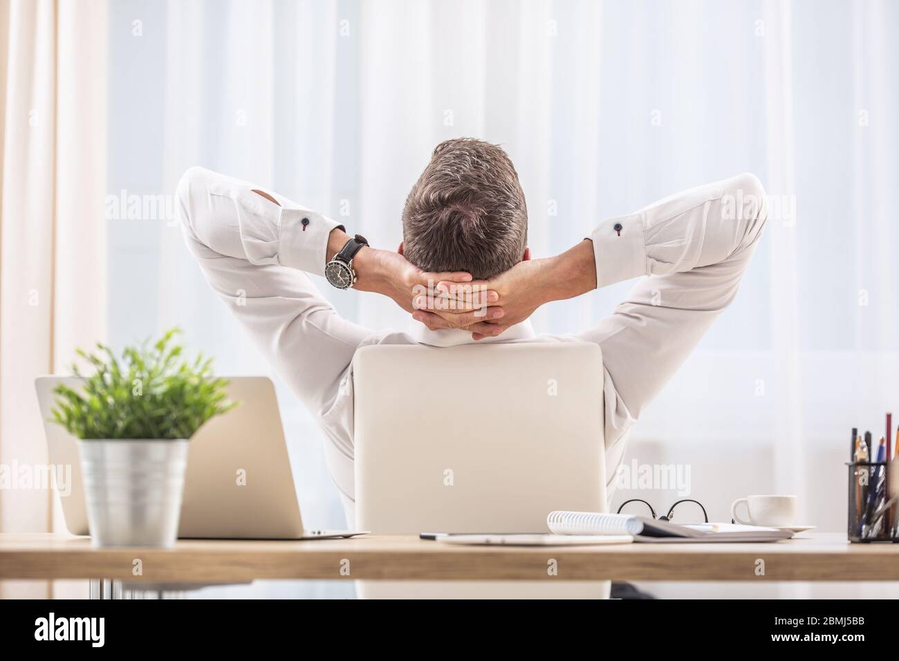 Geschäftsmann sitzt auf seinem Bürostuhl mit den Händen hinter dem Kopf, dreht den Rücken zum Schreibtisch und entspannt. Stockfoto