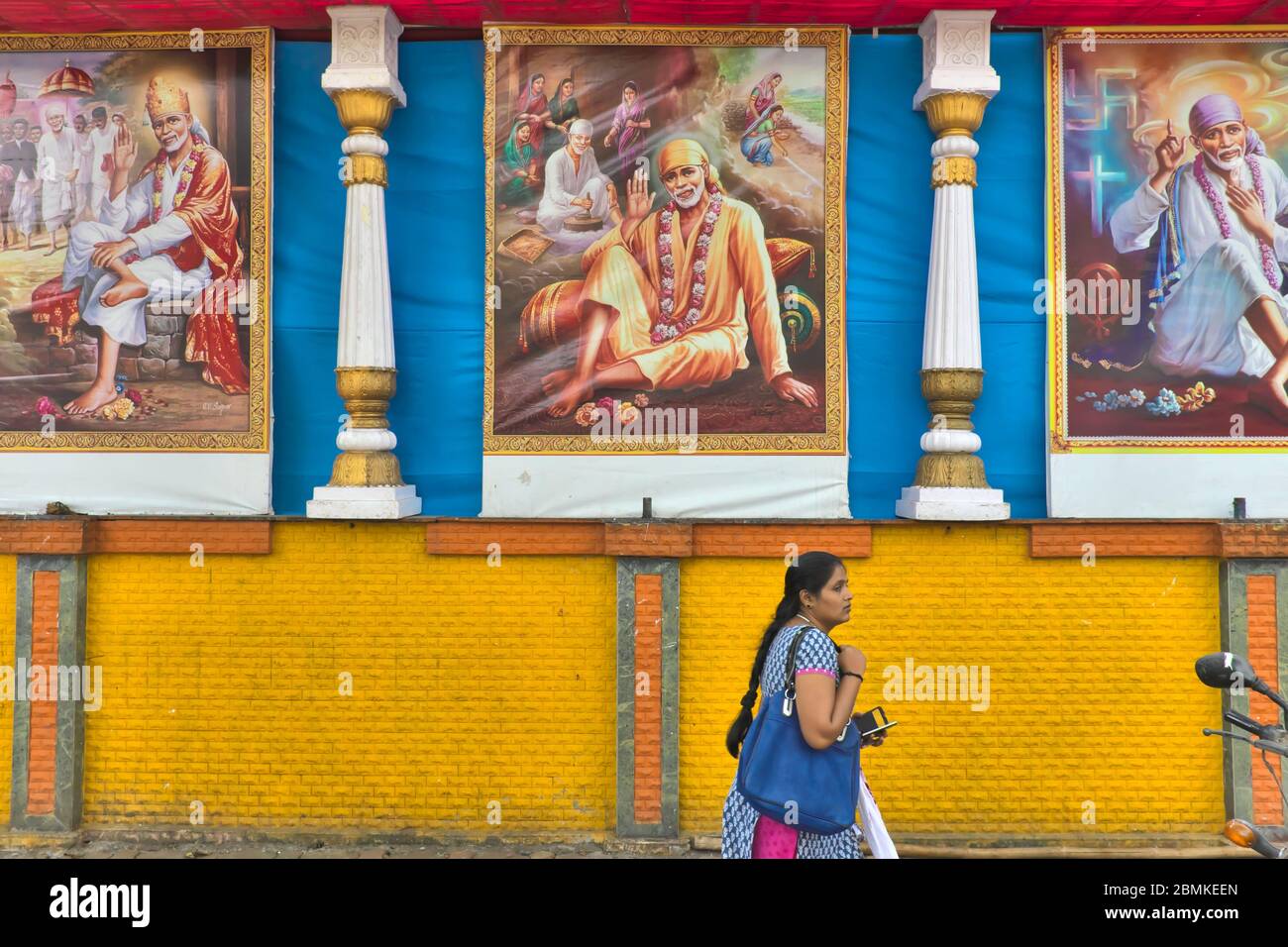 Eine Frau in Mumbai, Maharashtra, Indien, passiert eine blau-gelbe Wand mit drei Wandmalereien des Hindu-heiligen Sai Baba von Shirdi (1838-1918) Stockfoto