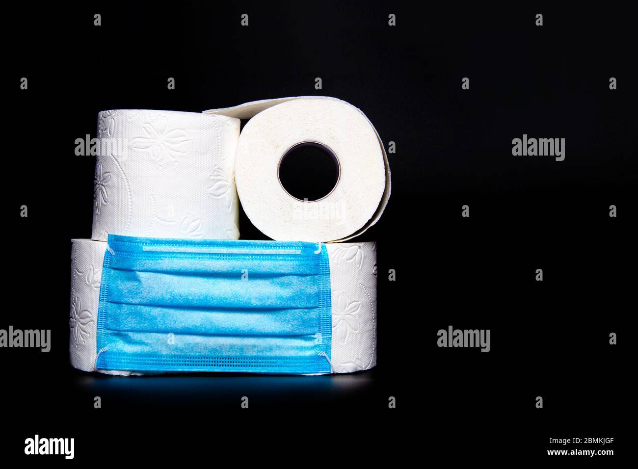 Rollen von weißem Toilettenpapier mit einer blauen medizinischen Gesichtsmaske sind auf einem schwarzen Hintergrund. Coronavirus Ausbruch Panik Konzept Stockfoto