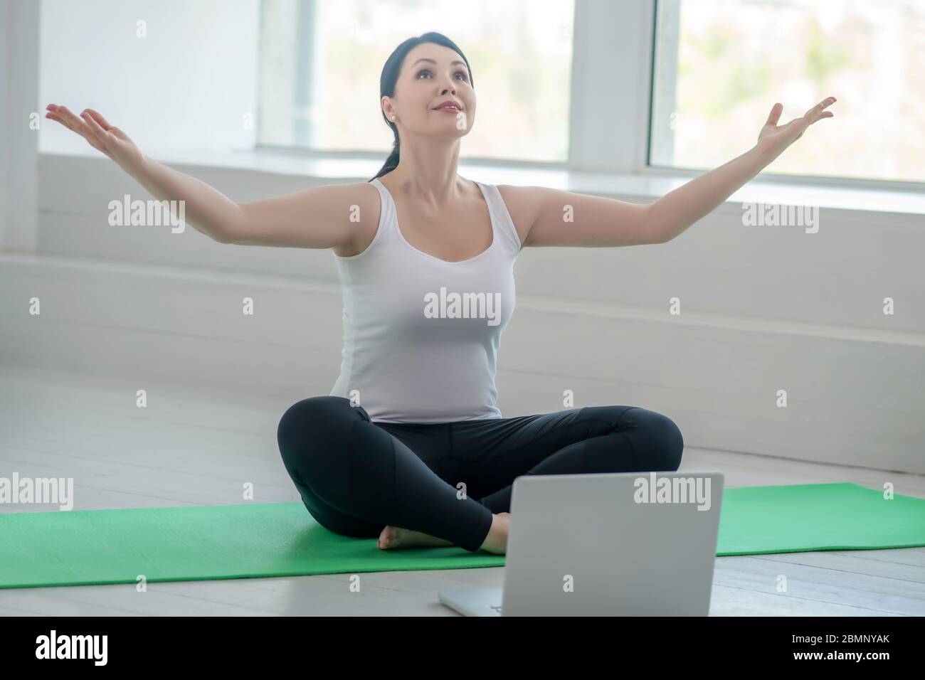Brünette Frau sitzt auf Teppich mit erhobenen Händen mit Online-Yoga-Lektion Stockfoto