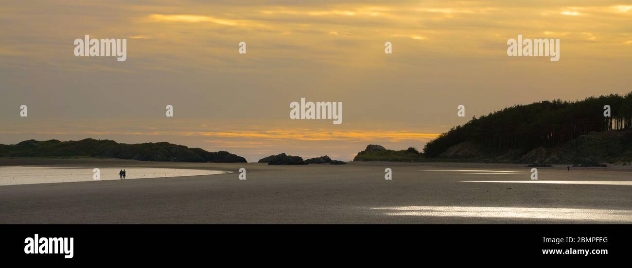 Panorama der Landschaft des britischen Strandes bei Sonnenuntergang. Ein Paar in Silhouette am Abend am Strand spazieren. Llanddwyn Island, Wales. Konzept Ruhe, Isolation. Stockfoto