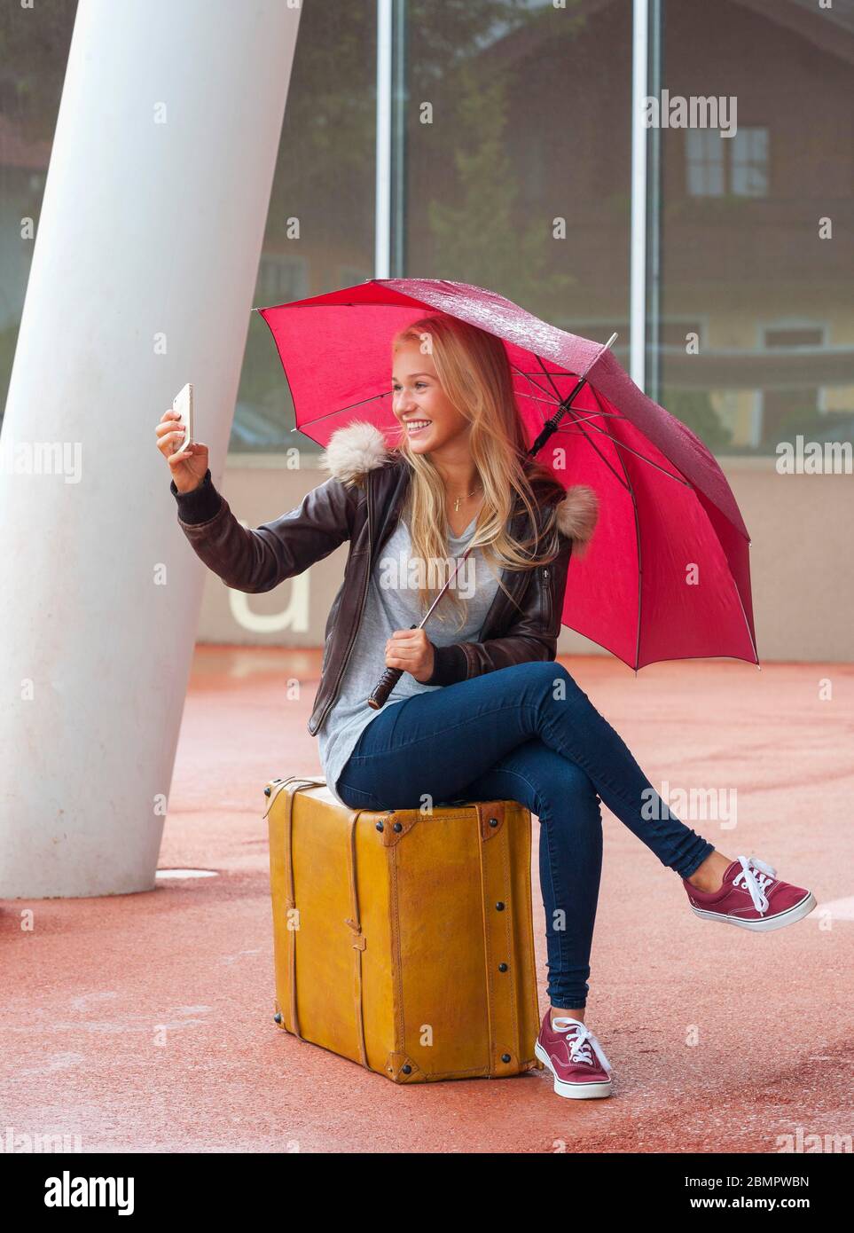 Mädchen mit rotem Schirm auf einem Koffer sitzend und Selfie machend, 16 Jahre, Österreich Stockfoto