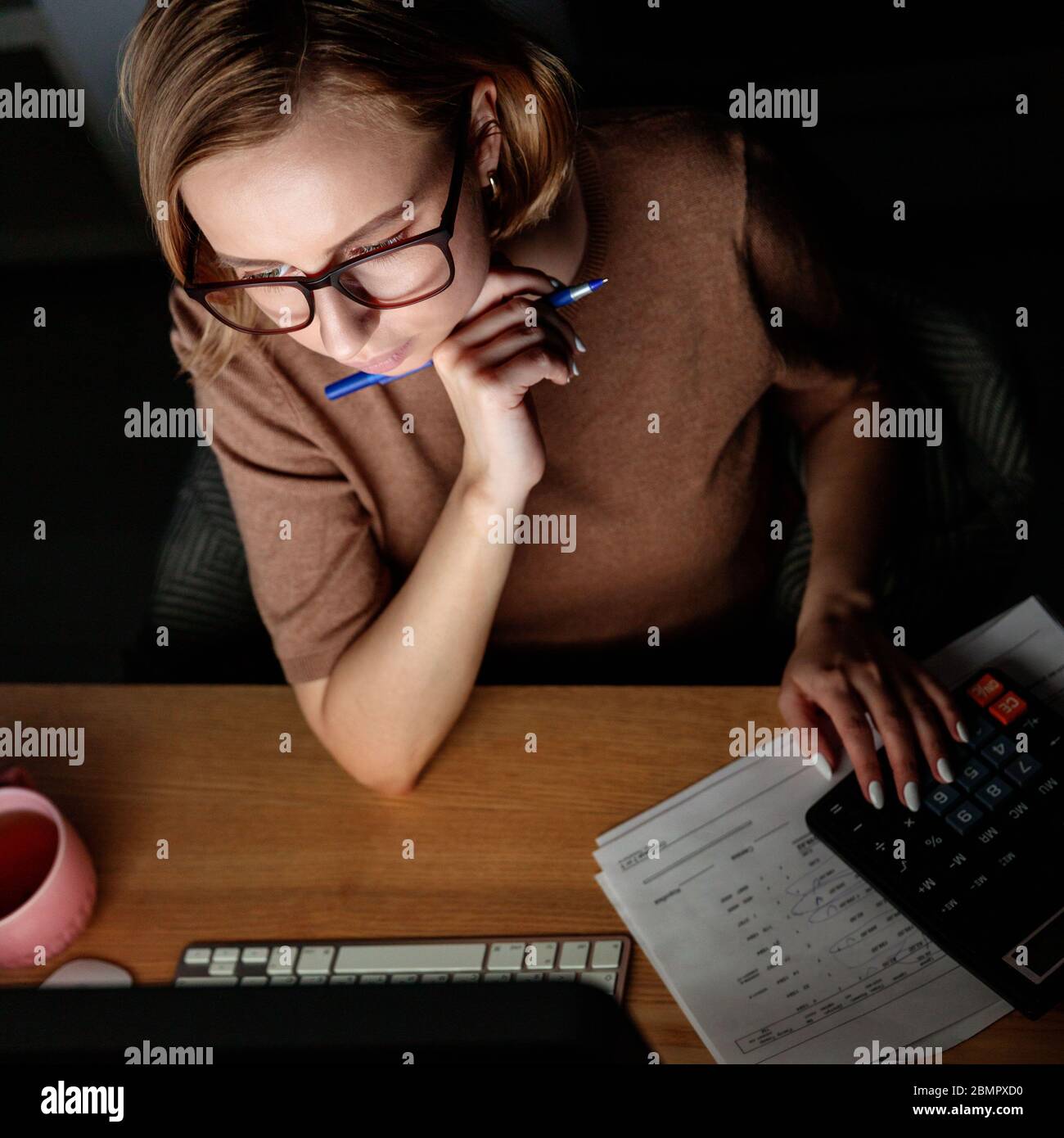 Frau in Brille mit Rechner zur Berechnung der Rechnung, Planung Kosten, halten Sie einen Stift in der Nähe Gesicht, die Arbeit auf Desktop-pc spät in der Nacht zu Hause Stockfoto