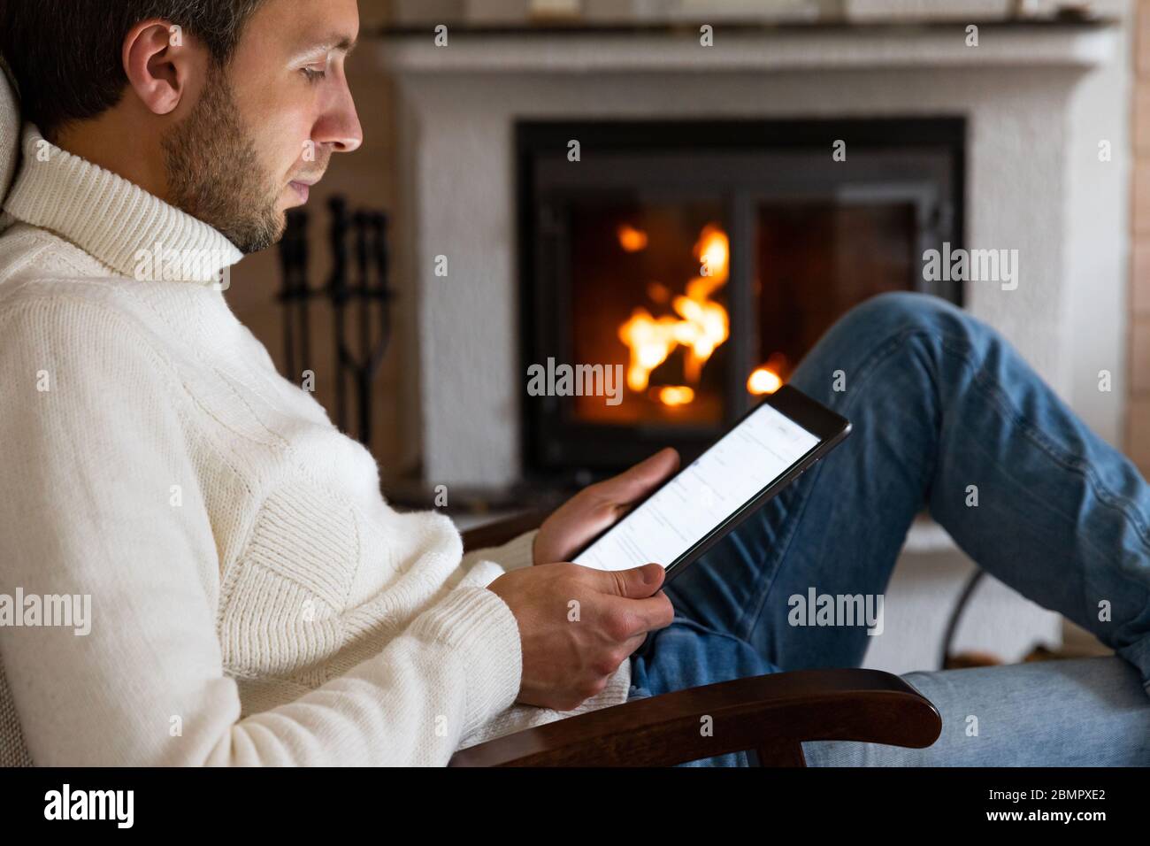 Nahaufnahme von kaukasischen Mann tragen hohen Hals weißen Pullover mit einem digitalen Tablet, Nachrichten lesen, scrollen sozialen Netzwerken, in einem Wohnzimmer durch die ausgeruht Stockfoto