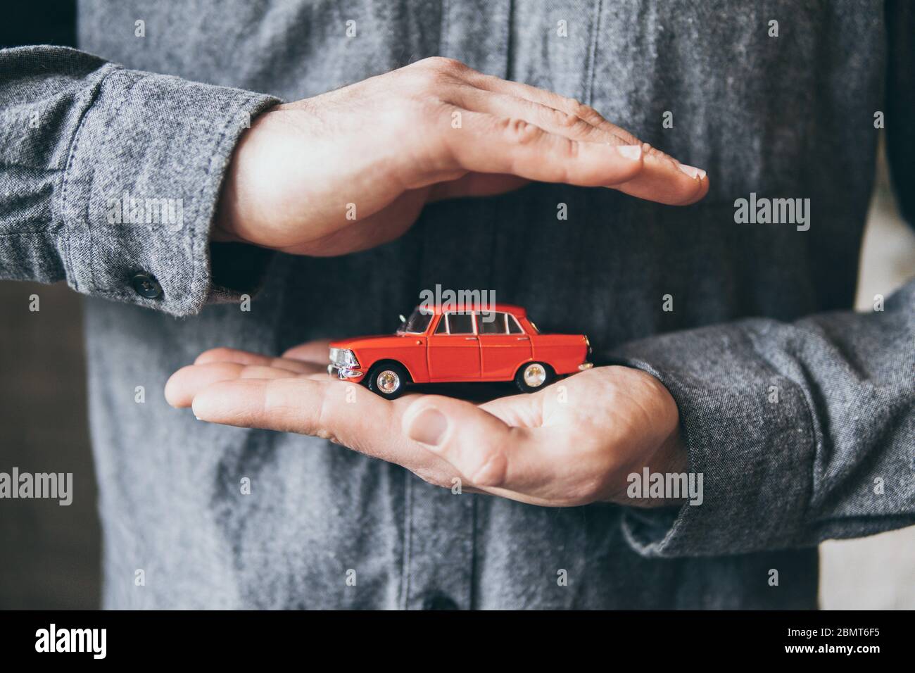 Männliche Hände halten und schützen ein rotes Spielzeugauto. Konzeptionelles Bild von Versicherung und Verkehrssicherheit. Stockfoto