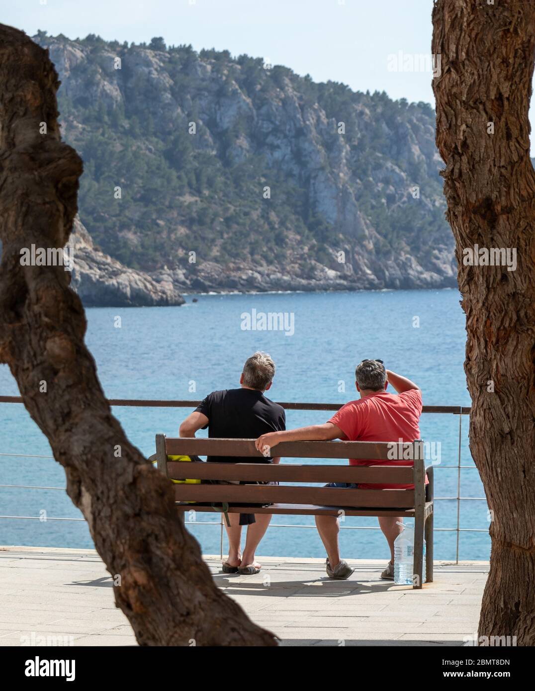 Sant Elm, Mallorca, Spanien - 12. April 2019: 2 Mann auf einer Holzbank unter einem großen Baum mit Blick auf die Küste von sant Elm, mallorca, spanien Stockfoto