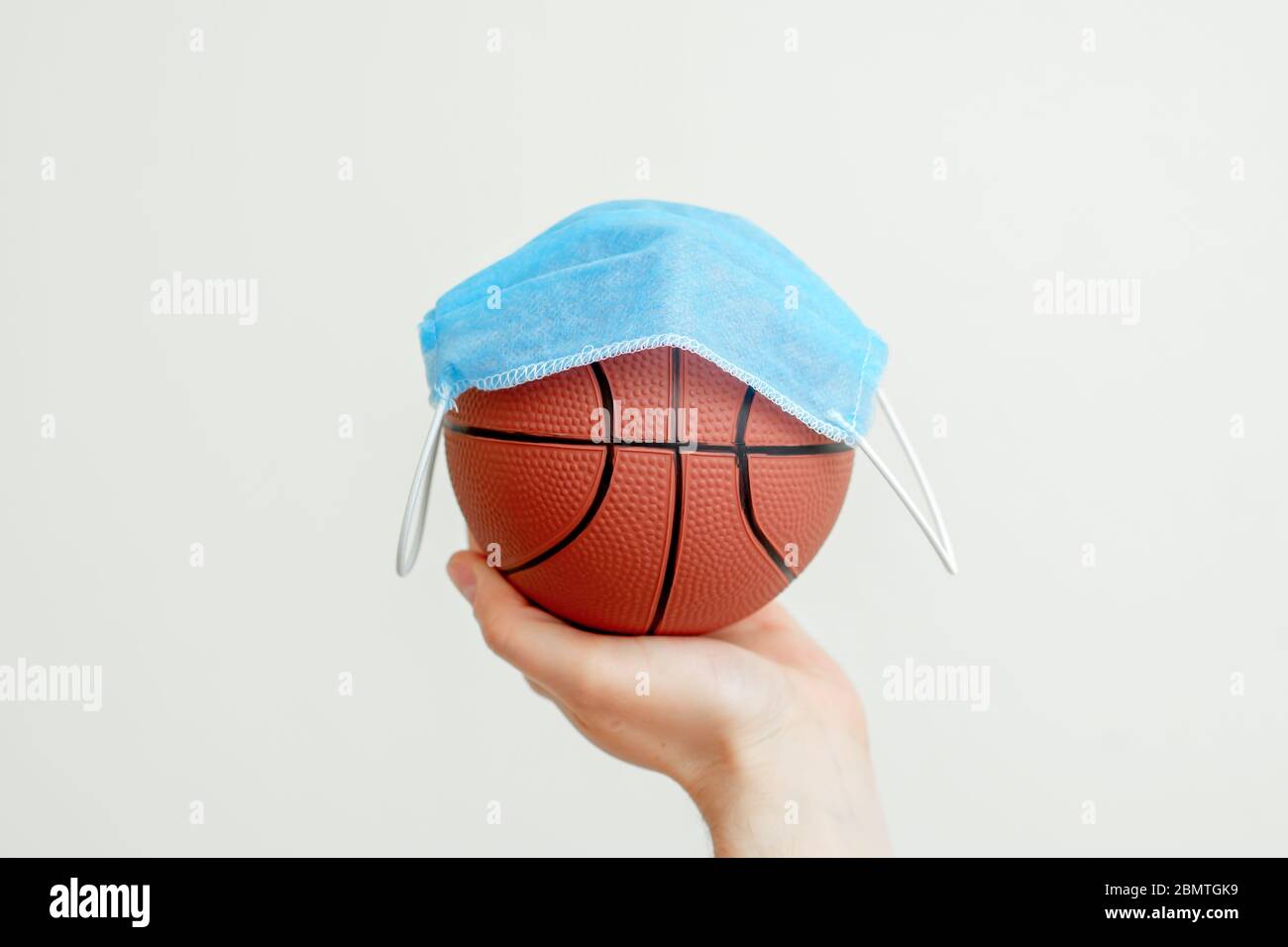 Hand des Mannes, der Basketball mit medizinischer Maske auf hellem Hintergrund hält. Stornierung von Basketballwettbewerben. Stockfoto