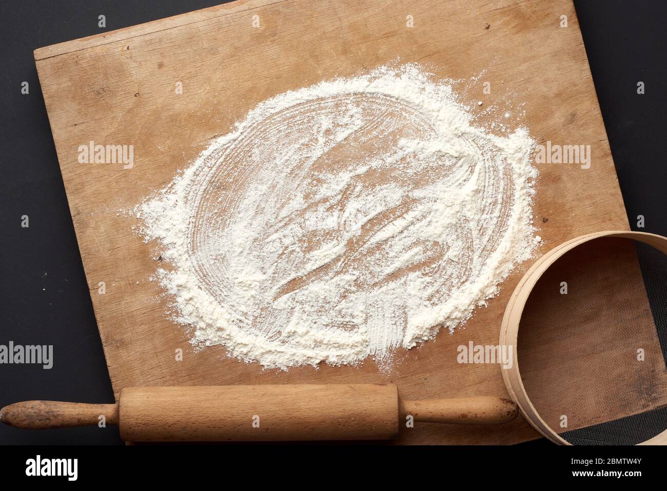 Weißes Weizenmehl verstreut und ein Holz-Nudelholz, rundes Sieb auf einem schwarzen Tisch, Draufsicht Stockfoto