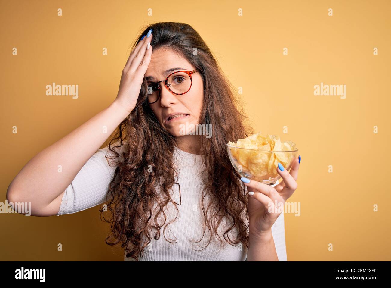 Junge schöne Frau mit lockigen Haaren hält Schüssel Chips Kartoffeln über gelbem Hintergrund gestresst mit Hand auf den Kopf, schockiert von Scham und Surpris Stockfoto