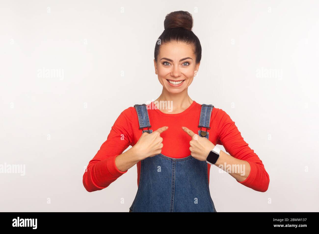 Das bin ich! Porträt von charmanten positive stilvolle Mädchen mit Haar bun in Overalls zeigt sich, stolz auf ihre Leistungen und Erfolg, Smilie Stockfoto