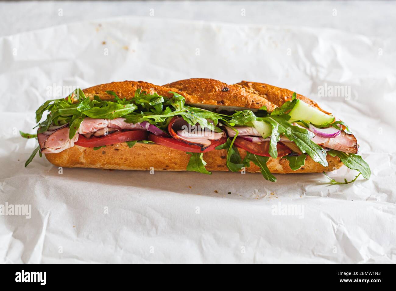 Baguette-Sandwich mit Fleisch, Tomaten, Gurken und Rucola auf weißem Papier. Essen zum Mitnehmen Konzept. Stockfoto