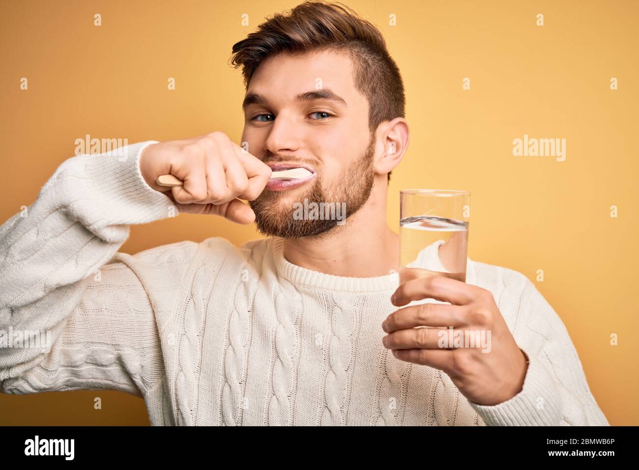 Junger gutaussehender kaukasischer Mann putzt seine Zähne mit Zahnbürste, Mundpasta und Glas Wasser, putzt Zähne und Zunge als gesunde Gesundheitsversorgung morn Stockfoto