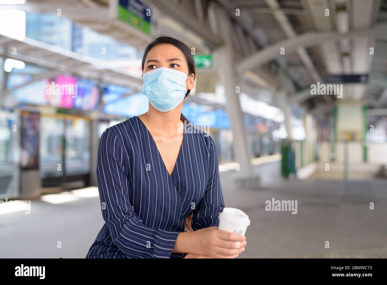 Junge asiatische Geschäftsfrau mit Maske Kaffee auf dem Weg während der Wartezeit am Skytrain-Bahnhof Stockfoto