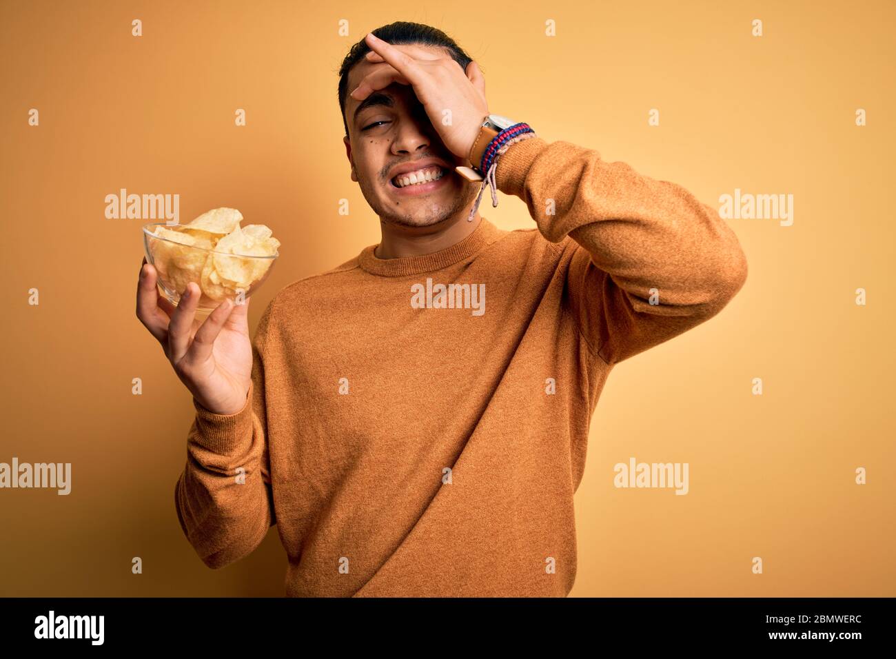 Junger brasilianer hält Schüssel mit Chips Kartoffeln über isoliert gelben Hintergrund gestresst mit Hand auf Kopf, schockiert mit Scham und Überraschung Gesicht, Stockfoto