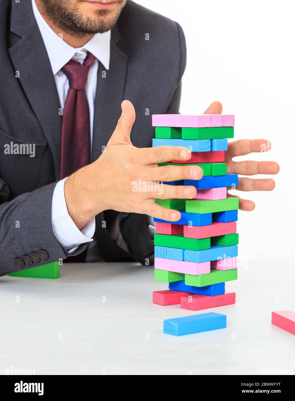 Bunte Blöcke ragen auf einem weißen Schreibtisch. Mann in Anzug versucht, das Gleichgewicht zu halten. Geschäftsstrategie, Risiko, Baukonzept Stockfoto