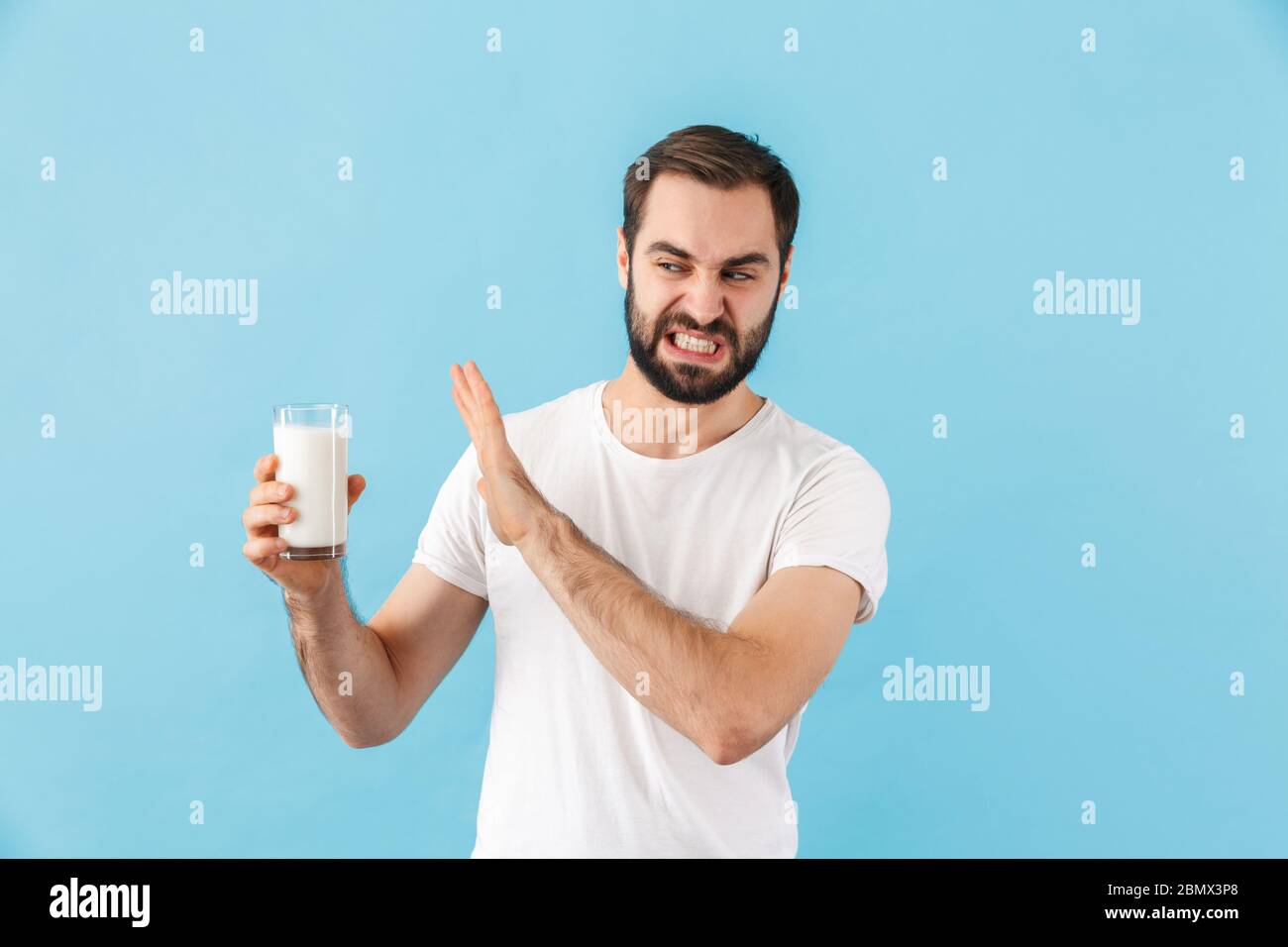 Porträt eines jungen angewiderten bärtigen Mannes trägt T-Shirt isoliert auf blauem Hintergrund, hält Glas Milch Stockfoto