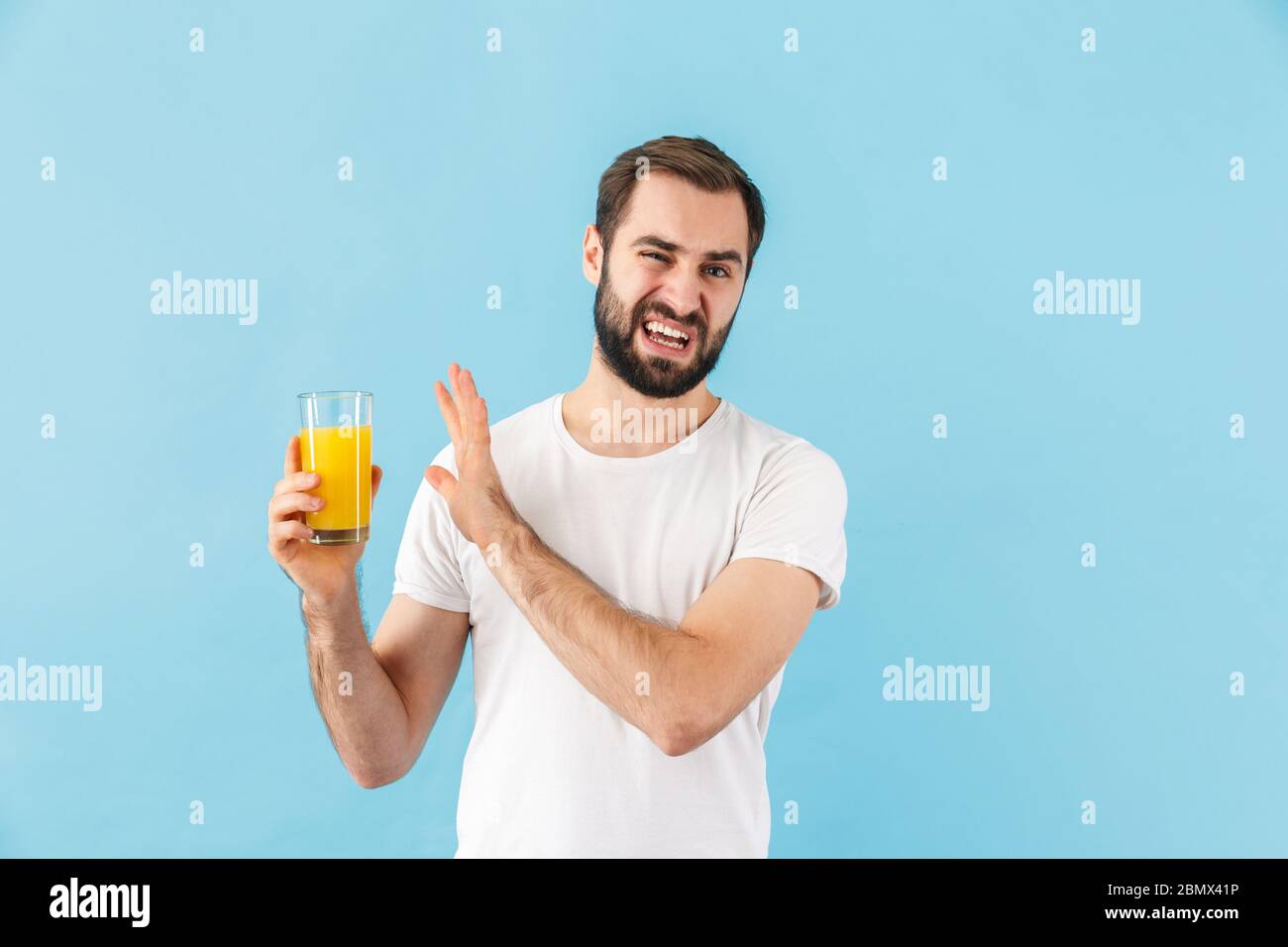 Schöner enttäuschter junger bärtiger Mann, der ein T-Shirt trägt, das isoliert auf blauem Hintergrund steht und ein Glas frischen Orangensaft zeigt Stockfoto