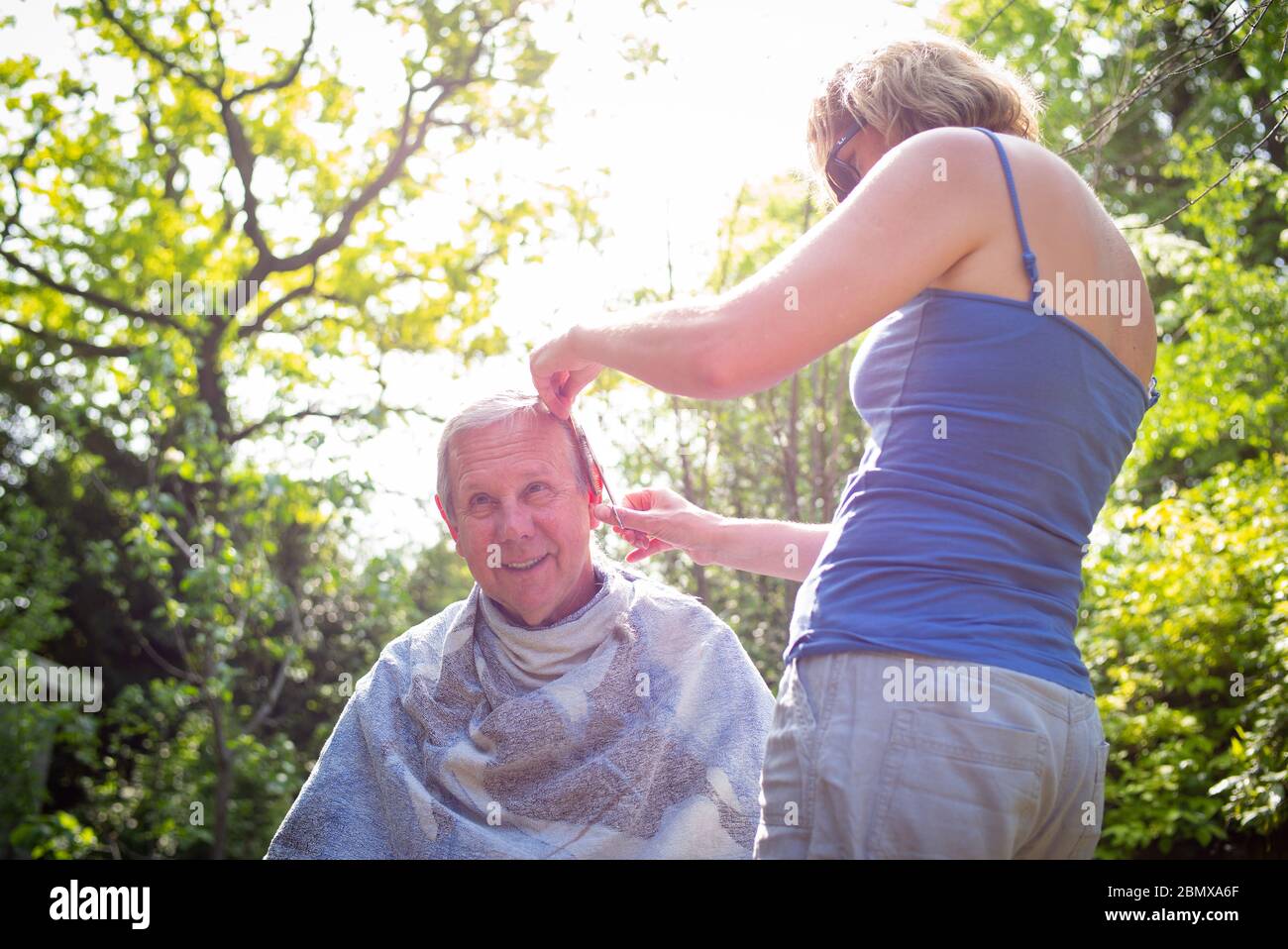 Die isolierende Schwiegertochter gibt ihrem Schwiegervater während der Sperrung im Garten während der Coronavirus-Pandemie einen Haarschnitt. Stockfoto