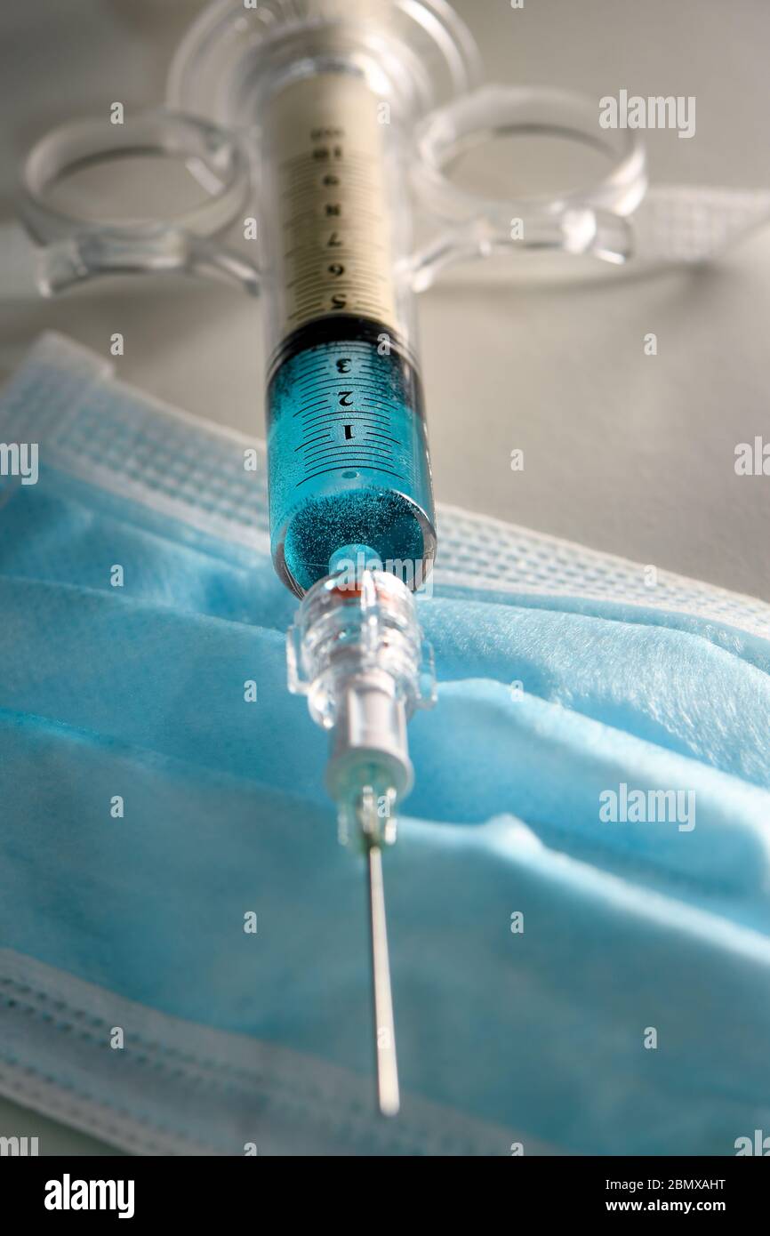Konzeptbild einer Spritze mit blauer Flüssigkeit auf einer blauen PSA-Gesichtsmaske. Könnte zur Veranschaulichung der COVID-19-Pandemie verwendet werden. Stockfoto