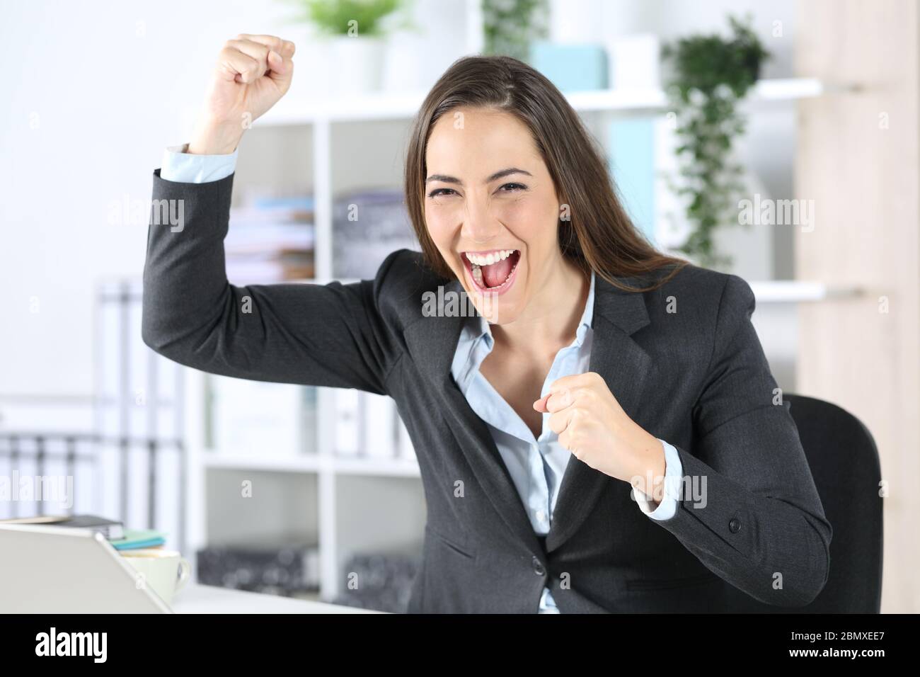 Eine begeisterte Geschäftsführerin feiert gute Nachrichten, wenn sie die Kamera im Büro auf ihrem Schreibtisch sieht Stockfoto
