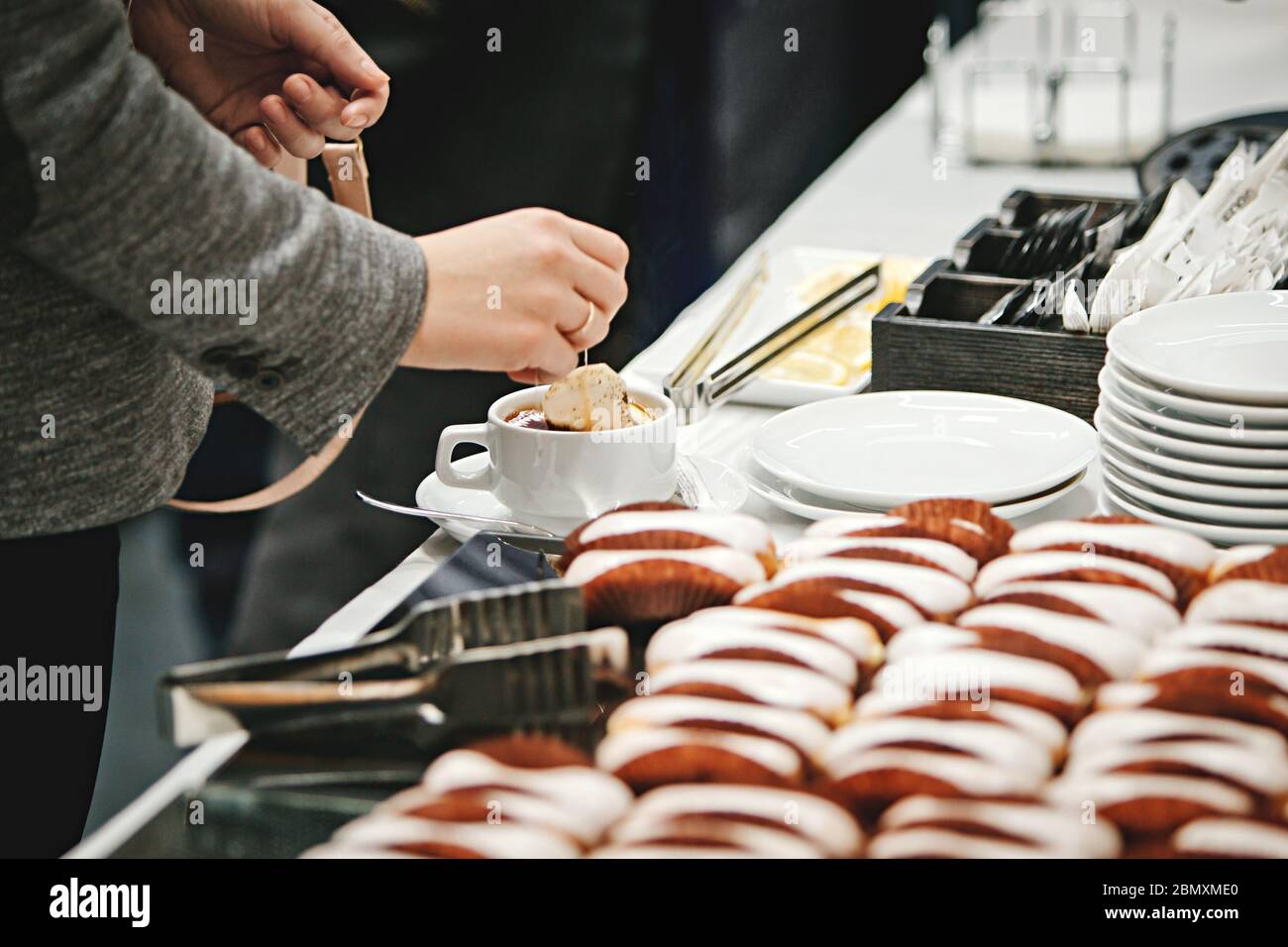 Eine Frau in einer grauen Jacke, deren Gesicht nicht zu sehen ist, Braut Tee in einer weißen Tasse auf dem Banketttisch, auf dem gebrühten Kuchen und einem Stapel weißer Platte liegen Stockfoto