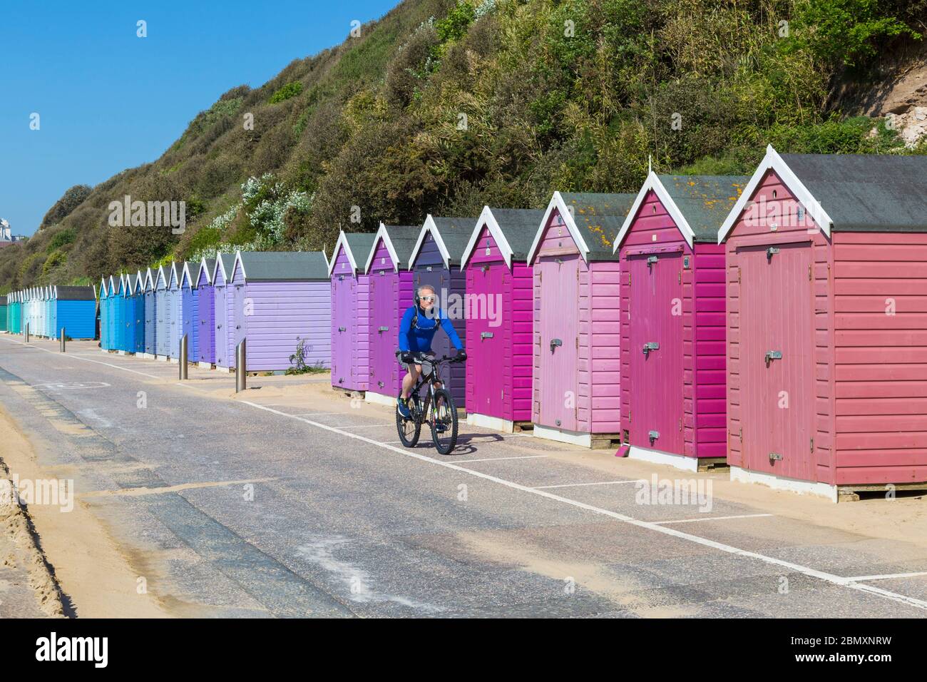 Radsportler, die an der Promenade entlang an bunten Strandhütten mit blauem Himmel an warmen sonnigen Tagen in Bournemouth, Dorset UK im Mai - Fahrrad fahren Stockfoto