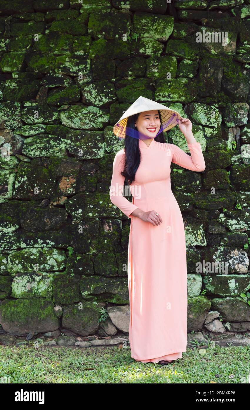 Eine Vietnamesin, die ein rosa Ao Dai Kleid trägt und vor den moosigen Mauern eines Tempels in Hue City lächelt Stockfoto