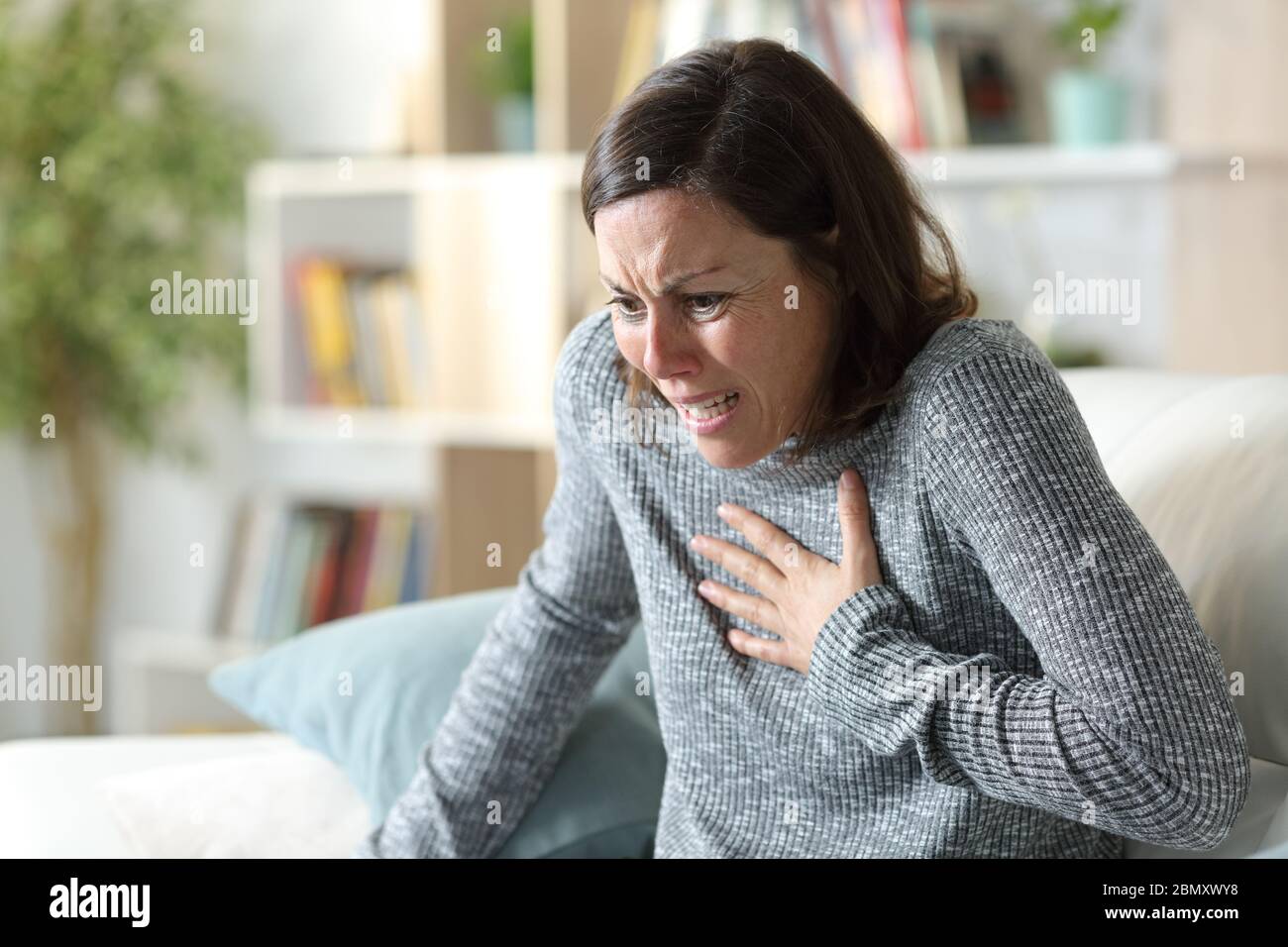 Verängstigte Erwachsene Frau, die einen Herzinfarkt hat und die Brust schmerzt, sitzt zu Hause auf einer Couch Stockfoto