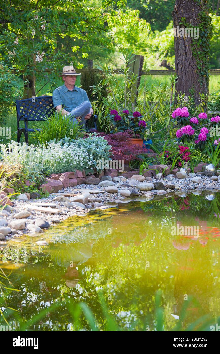 Reifer Mann sitzt auf einer Bank an einem Teich in einem Garten mit bunten Blumen - konzentrieren Sie sich auf den Mann Stockfoto