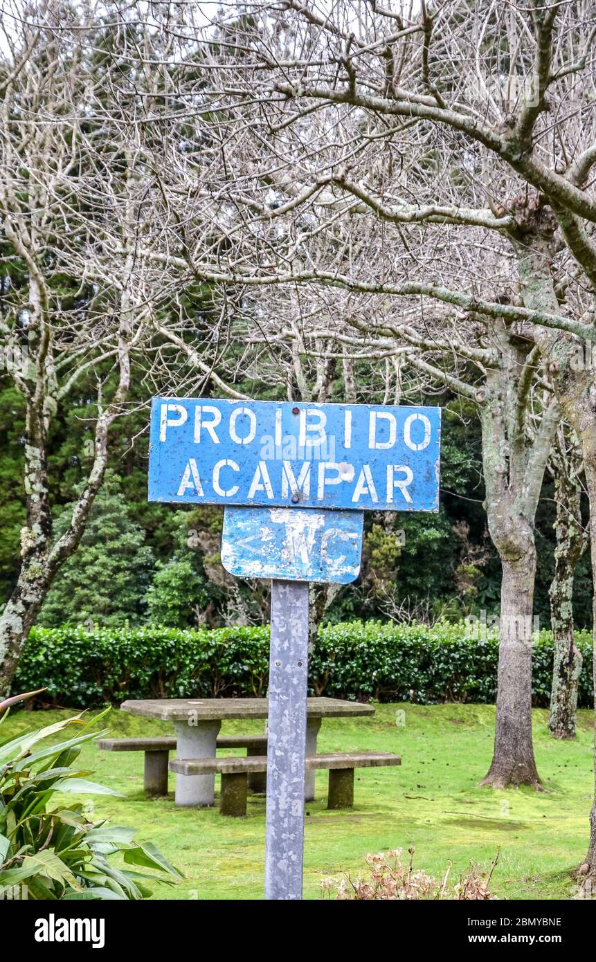 Altes blaues Schild mit Text Proibido Acampar in portugiesischer Sprache. ÜBERSETZUNG: Camping verboten. Park mit grünen Bäumen, Bänken und Tischen im Hintergrund. Kein Campingschild, keine Übernachtung. Stockfoto