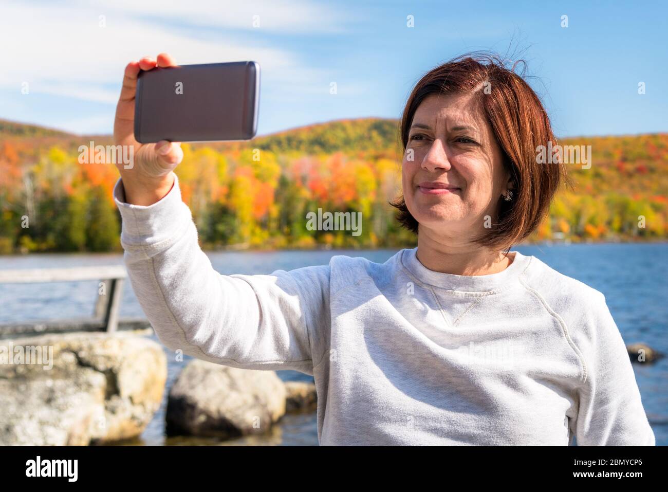 Eine Touristenin, die an einem sonnigen Tag ein Selfie am Ufer eines Sees, umgeben von Wäldern, auf dem Gipfel des Herbstes, abnimmt Stockfoto