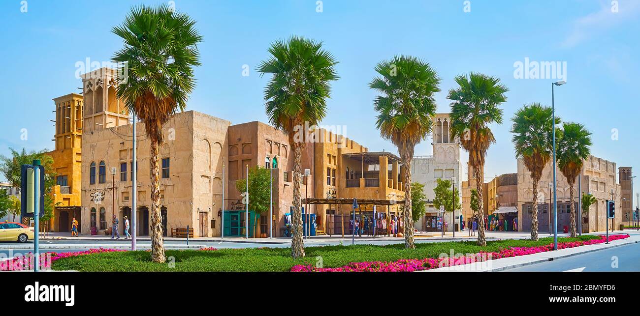 DUBAI, VAE - 2. MÄRZ 2020: Panorama der Al Seef Straße mit üppigen Palmen, Blumenbeeten und historischen Lehmbauten des Heritage Village mit barjeel Windc Stockfoto