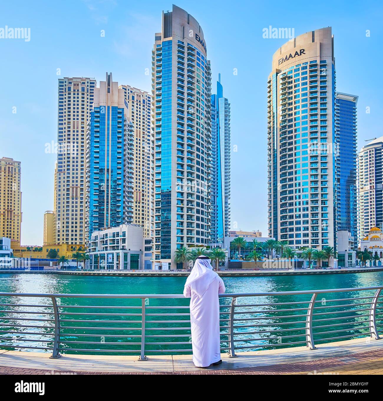 DUBAI, VAE - 2. MÄRZ 2020: Der Emirati-Mann in traditioneller weißer Kleidung (Kandura und Ghutra) steht an der Küste der Dubai Marina und beobachtet moderne BU Stockfoto