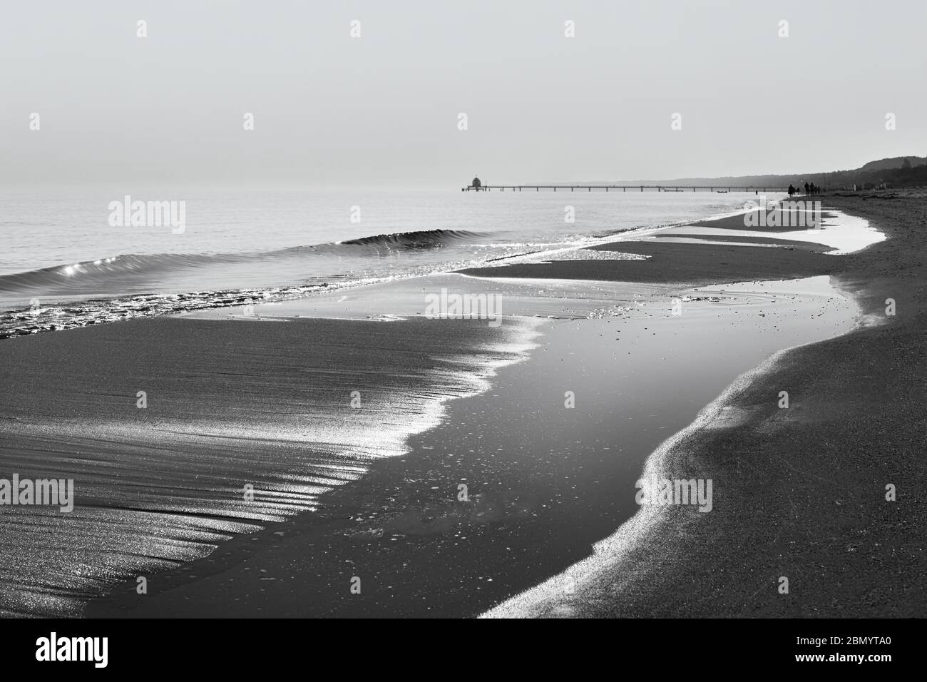 Blick entlang der Wasserlinie mit Welle, Wasserflächen und Spaziergängern sowie einem Pier im Hintergrund - Bild in Schwarz-Weiß - Ort: Ostsee Stockfoto